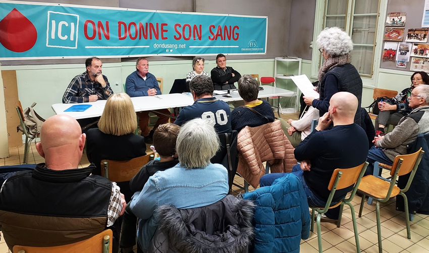 Lors de l'assemblée du don du sang, une femme est début au milieu des participants assis et lit un texte. En face d'elle, se tiennent quatre personnes assises derrière une table, parmi lesquelles se trouvent le Maire et la Présidente de l'association.